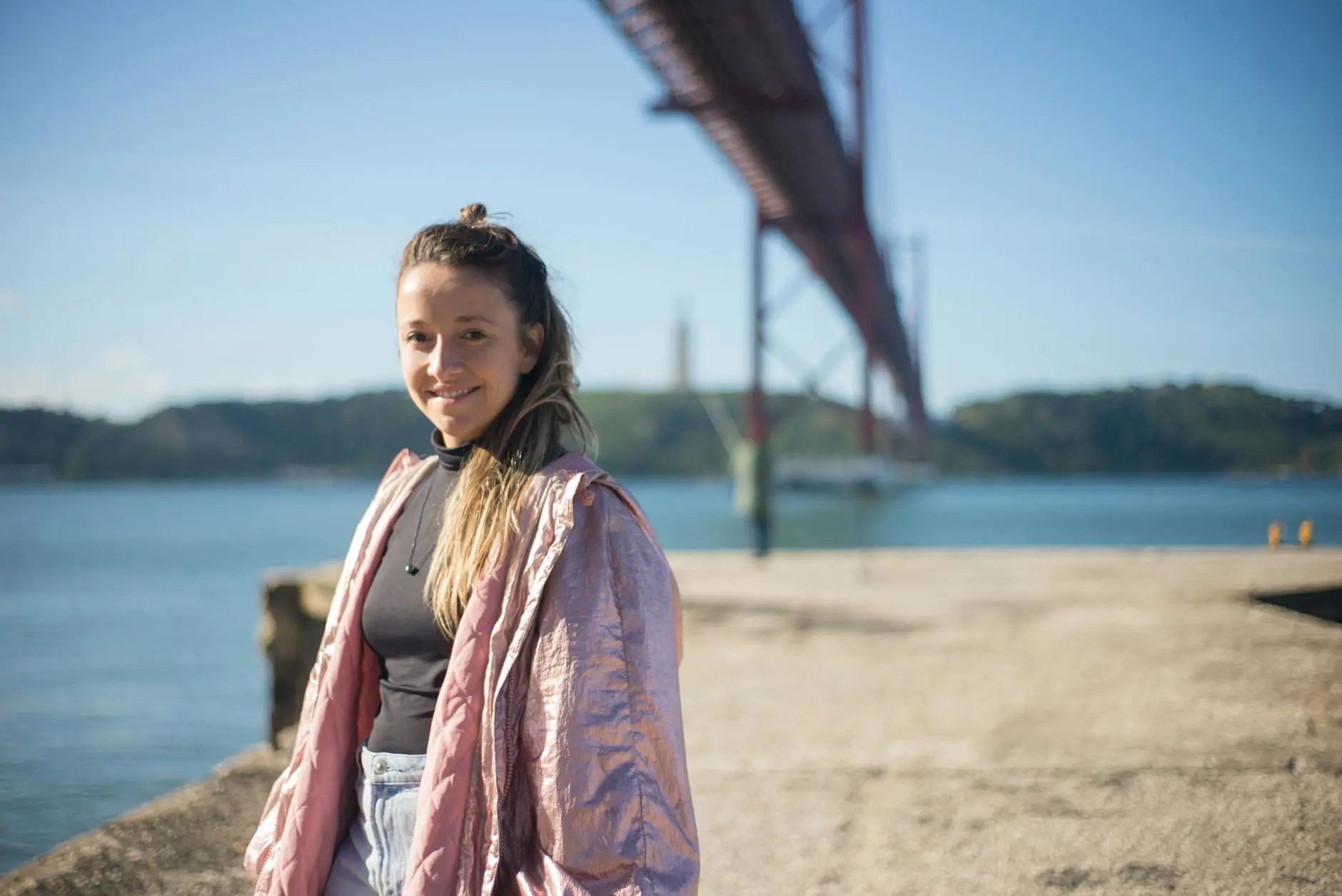 A young woman smiles with the 25 de Abril Bridge in Lisbon, Portugal in the background.